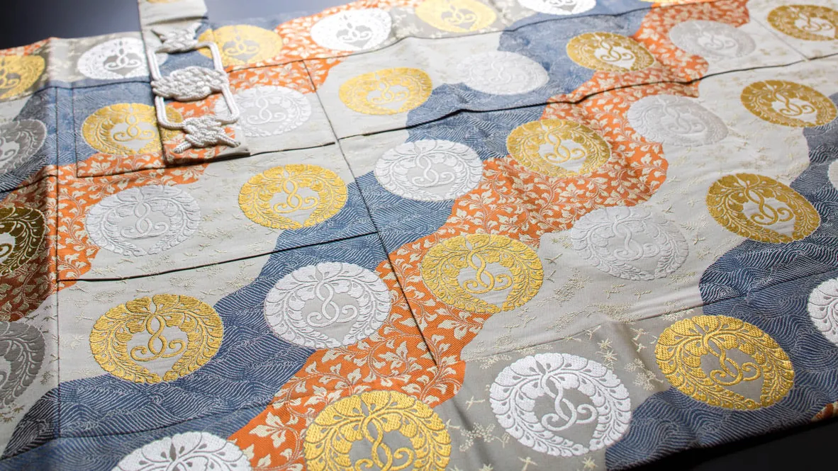 A five-strip kesa of orange, blue and grey brocade with white family-crest roundels and a clasp