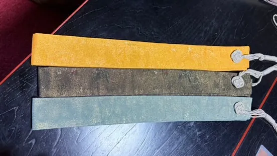 Three narrow washable strips in yellow, dark olive and grey-green lying on a dark table, each with a silver-grey tassel cord at one end