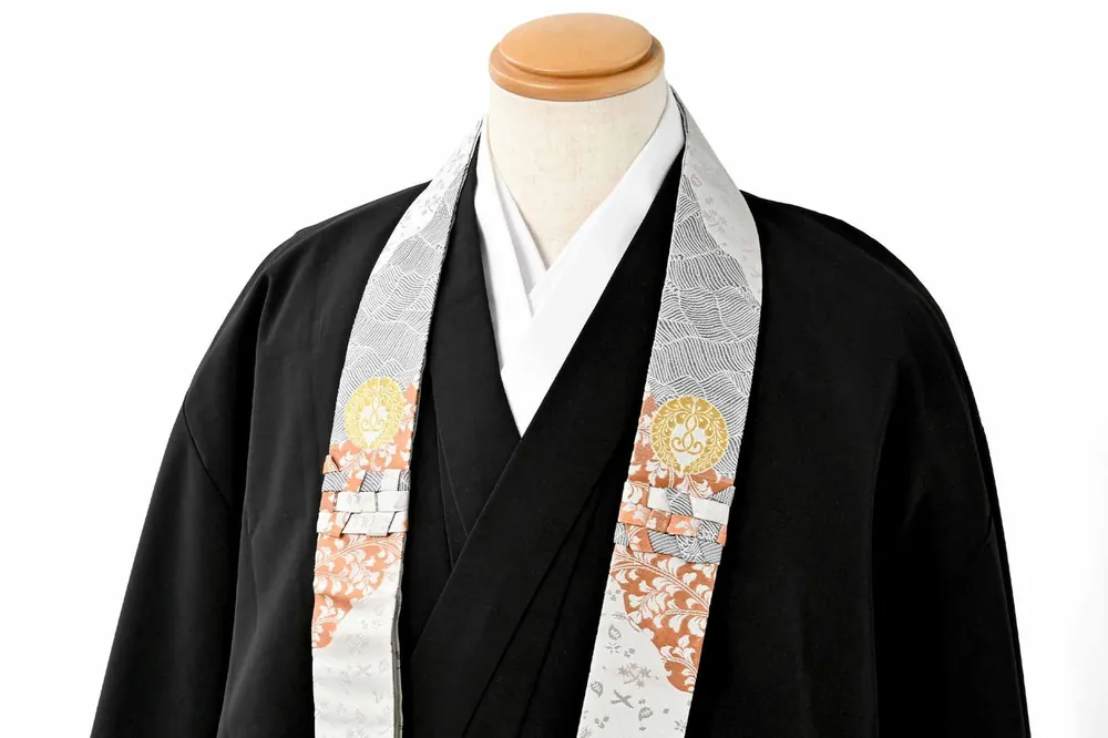 A black priest’s robe on a mannequin with a white and silver wagesa that has gold crests and a pink and orange floral patch on each end