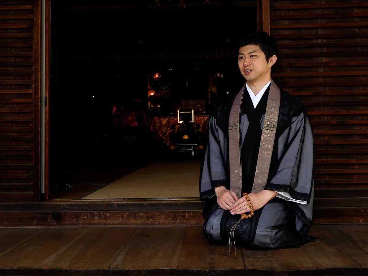 A man in a grey-brown sheer robe kneeling in a temple, wearing a dark washable wagesa and holding wooden prayer beads